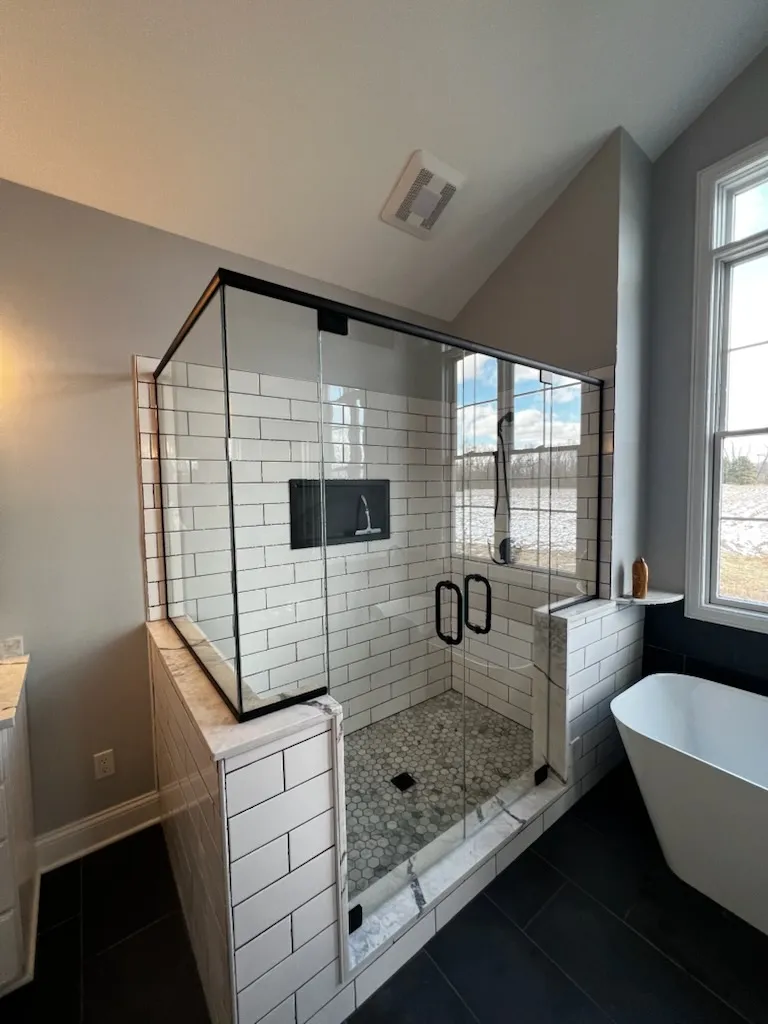 Glass shower enclosure with black trim and handles on a white subway tile shower beside a soaking tub.