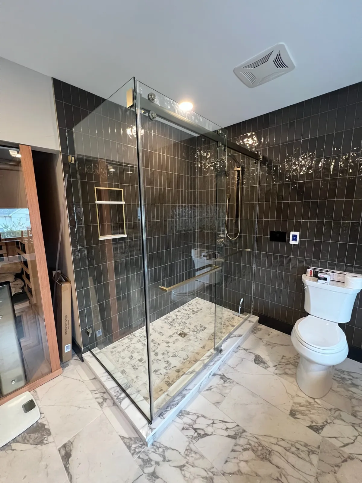 Corner sliding glass shower enclosure with gold towel bar against dark vertical tile.
