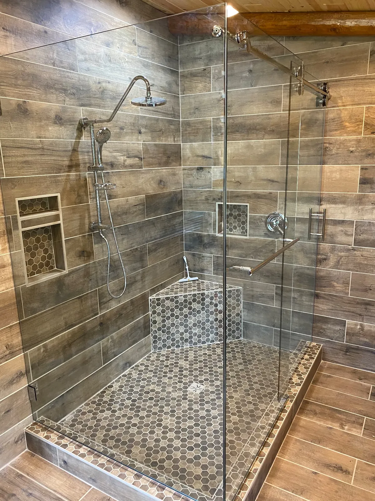 Frameless sliding glass shower door on a chrome rail with wood-look tile and a corner seat.